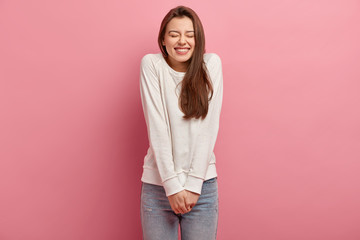 Funny overemotive woman keeps hands together, laughs at comic story, keeps eyes closed, dressed in casual long sleeve sweater and denim trousers, isolated over pink background, has good time