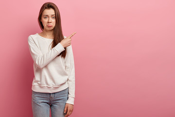 Frowned unhappy lady purses lower lip, directs at empty space, wears casual sweatshirt and jeans,...