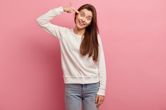 Happy young woman shoots with fingers in temple, makes suicide gesture, smiles broadly, tilts head, tries kill herself, wears jumper and jeans, isolated over pink background, fed up with something