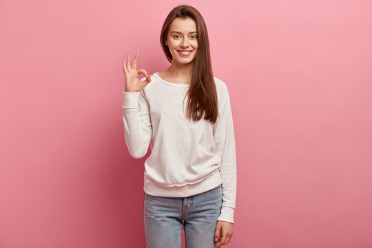 Indoor shot of good looking brunette woman customer shows great sign, satisfied with good quality of product, dressed in casual clothes, models against pink background. Girl demonstrates zero sign