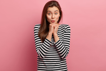 Photo of attractive woman has green eyes, pouts lips, keeps hands under chin, feels delighted, wears casual striped jumper, looks directly at camera, isolated over pink background. Facial expressions