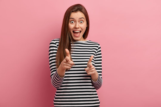 Emotional pleased young female model makes finger gun gesture directly at camera, openes mouth with amazement, wears striped clothes, isolated over pink background, greets friend, gestures indoor