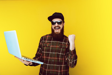 Excited bearded man in sunglasses and hat, holding laptop and celebrating fist up