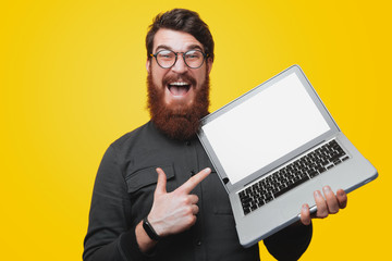 Happy bearded man in glasses holding and pointintg at laptop while looking at camera