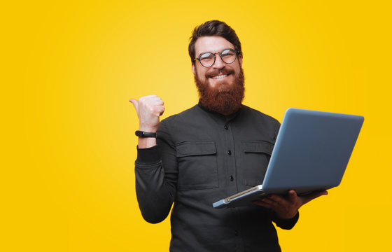 Cheerful Bearded Man Is Holding Laptop And Poiting Bkack, Looking At Camera Over Yellow Background