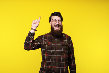 A cheerful bearded man in round glasses is pointing with finger up while looking at camera