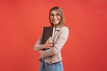 Young smiling student or intern in eyeglasses standing with a folder on red background. - obrazy, fototapety, plakaty
