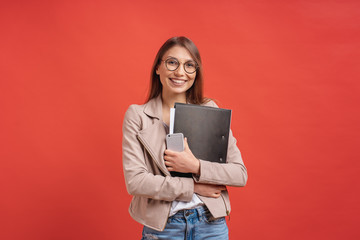 Young smiling student or intern in eyeglasses standing with a folder on red background. - obrazy, fototapety, plakaty