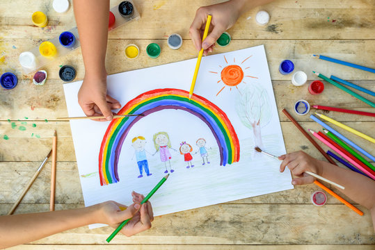 Happy family concept. Co-creation. Children hands draw on a sheet of paper: father, mother, boy and girl hold hands against background of rainbow and sunny sky. Close-up.