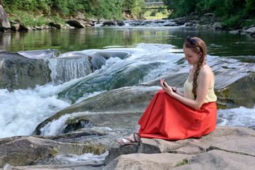 Beautiful long-haired girl in a red skirt with a smartphone sitting on a rock on a background of...