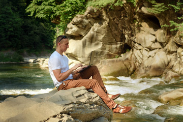 Work in nature concept. Man with notebook sitting at the bank of river over the waterfall and green trees. Side view