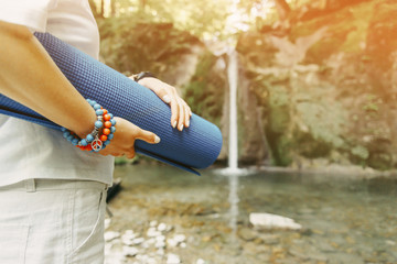 Woman with yoga mat standing in front of waterfall.