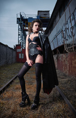 Fashion shot: portrait of beautiful goth girl (informal model) in leather coat, stockings and corset standing at railroad (industrial area)