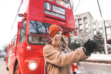 Cute positive girl with winter clothes standing near the red bus and cafe to take selfie on smartphone. Attractive girl walking along the streets of the winter town and take selfie.