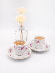 Two floral ornamented porcelain cups of espresso coffee and a glass vase with two synthetic chrysanthemum flowers isolated on a white background