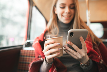 Fototapeta premium Happy girl uses a smartphone and drinks coffee in a cozy cafe. Close-up of a smartphone and a cup of coffee in their hands. Background. Copyspace