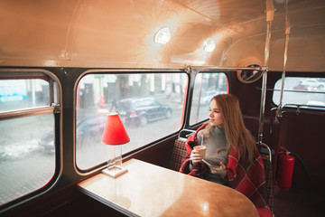 Fototapeta na wymiar Young attractive woman sitting at the table in the bus cafe is covered with a blanket, holds a cup of coffee in her hands and looks in the window. Beautiful girl sits in a cozy original cafe.