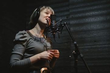 Naklejka premium Woman a singer in a headphones is performing a song in a sound recording studio and is standing in front of the microphone.