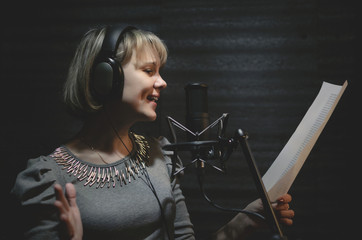 Woman a singer in a headphones is performing a song in a sound recording studio and is standing in front of the microphone.