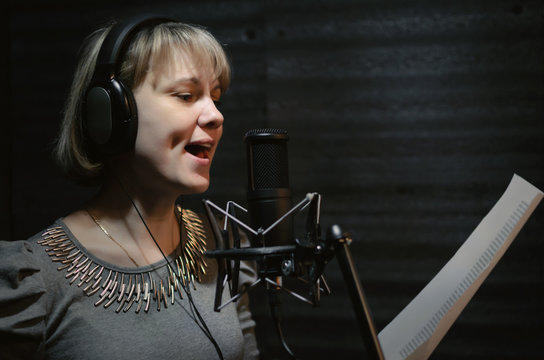 Woman a singer in a headphones is performing a song in a sound recording studio and is standing in front of the microphone.