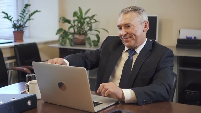 Senior entrepreneur in suit working on laptop computer in office. Mature ceo looking to camera and smiling on workplace in consulting company. Portrait. Slow motion.