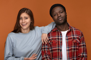 Portrait of stylish young dark skinned man having serious grumpy look being insulted with joke or prank made by his girlfriend who is laughing next to him, having clueless facial expression