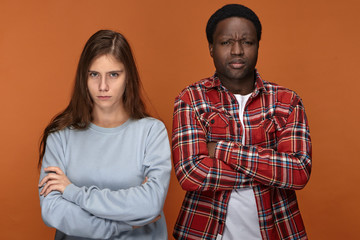 Stubborn young interracial couple having disagreement about plans on weekend looking at camera with grumpy facial expressions, keeping their arms crossed, frowning, not talking to each other