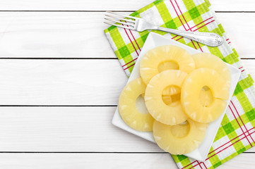 Plate with canned pineapple rings with fork and kitchen towel on white wooden table. Top view. Space for text.