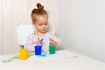 Beautiful little girl draws with finger paints on a white sheet of paper. Creative child development in kindergarten or free time at home