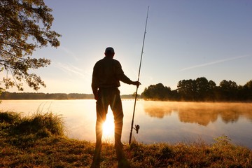 Silhouette of angler standing on the lake shore during misty sunrise
