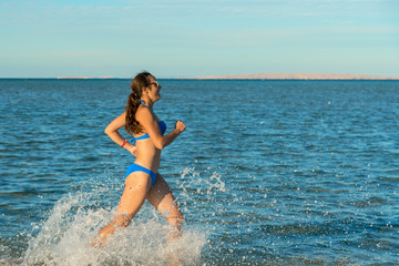 A sexy young brunette woman or girl wearing a bikini running through the surf on a deserted tropical beach with a blue sky. Young woman running by the sea.