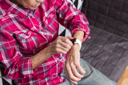 A very old senior Caucasian grandmother with gray hair and deep wrinkles sits at home on the couch in jeans and a red plaid shirt and uses a smart watch on her wrist. Pensioner and technology