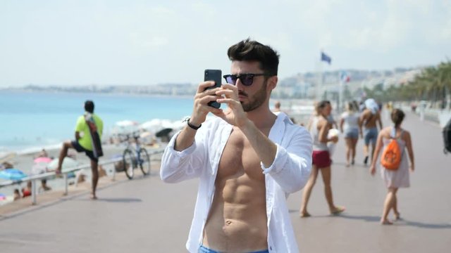 Young male tourist walking, exploring Nice in France, using cell phone to film video or take photos of the beach and seafront