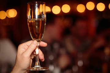 Champagne or white wine glass in woman hand at party or celebration of new year or birthday for toast cheers, blurred bokeh yellow rounds background, joy time rest, details and reflections warm light