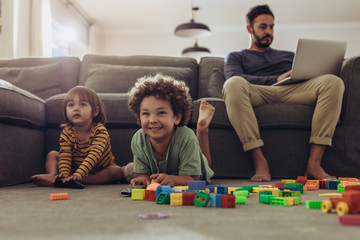 Man working on laptop sitting at home with kids playing on the f