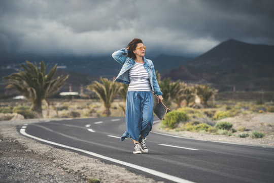 Middle aged woman skateboard smiles satisfied holding her dark hair with hand curved road with thunderstorm horizon striped knit long skirt sneakers wind free time summer holidays sunset sport