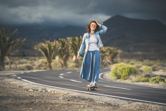 Middle aged woman with striped vest long skirt sneakers walking along road with skate temporal hand horizon keeps her hair blowing wind free time summer holidays sunset sport