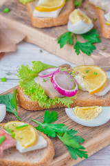 Sandwich with herring and tuna fillets with fresh green salad, onion ring, a quarter of eggs, home cheese, piece of lemon, micro greens and scallions on the wooden desk