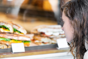 Closeup of woman looking at display of sandwiches behind glass in cafe lunch restaurant storefront