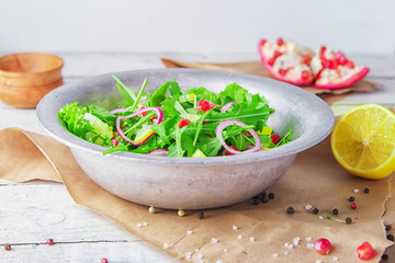 Fresh salad with pomegranate seeds, arugula, red onion, goat cheese, lemon slices, lettuce leaves, flavored with olive oil and balsamic dressing on a vintage metallic bowl