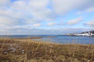 Russian Arctic View, North. Sky with clouds and blue sky reflection in water with waves and snow mountains background with stones grass foreground. Picturesque landscape view, nature scenery beauty
