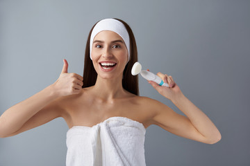 Cheerful young woman holding pore cleansing brush and showing thumbs up
