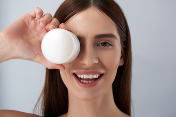 Charming brunette girl covering eye with white jar of cosmetic cream