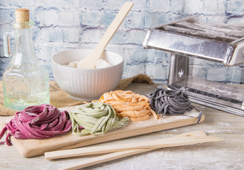 Variety of colored fresh raw uncooked homemade pasta tagliatelle green spinach, pink beetroot, orange carrots and black cuttlefish ink with flour on desk over old wooden table