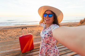 People, travelling and vacation concept - happy young woman with suitcase takes selfie portrait on the beach