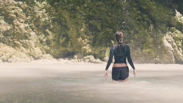 Traveling woman in mountain lake and water stream flowing from tropical waterfall. Tourist woman bathing in waterfall lake in rainforest. Girl on background waterflow from cliff in jungle.