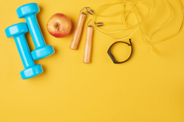 Fitness concept with blue dumbbells, skipping rope and fitness tracker on a yellow background, top view flat lay
