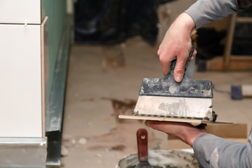 Closeup professional builder's hand smears cement glue on the ceramic tile with trowel to glue in bathroom. Concept renovation repair, overhaul, contract tiler, team of construction repairman