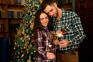 Merry Christmas and Happy New Year! Attractive young couple is celebrating holiday at home together, drinking champagne and smiling with Bengal lights in hand