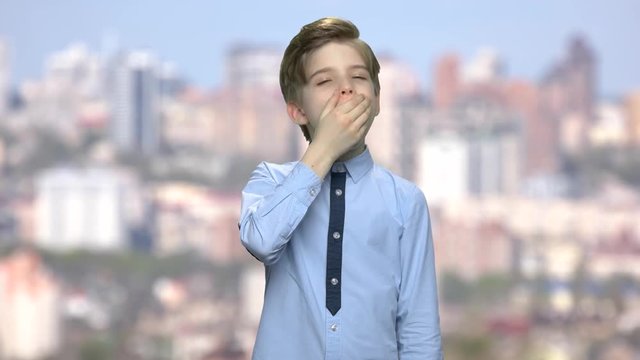 Tired young boy is yawning. Sleepy exhausted boy in blue sirt is yawning and covering mouth with hand. Blur city background.